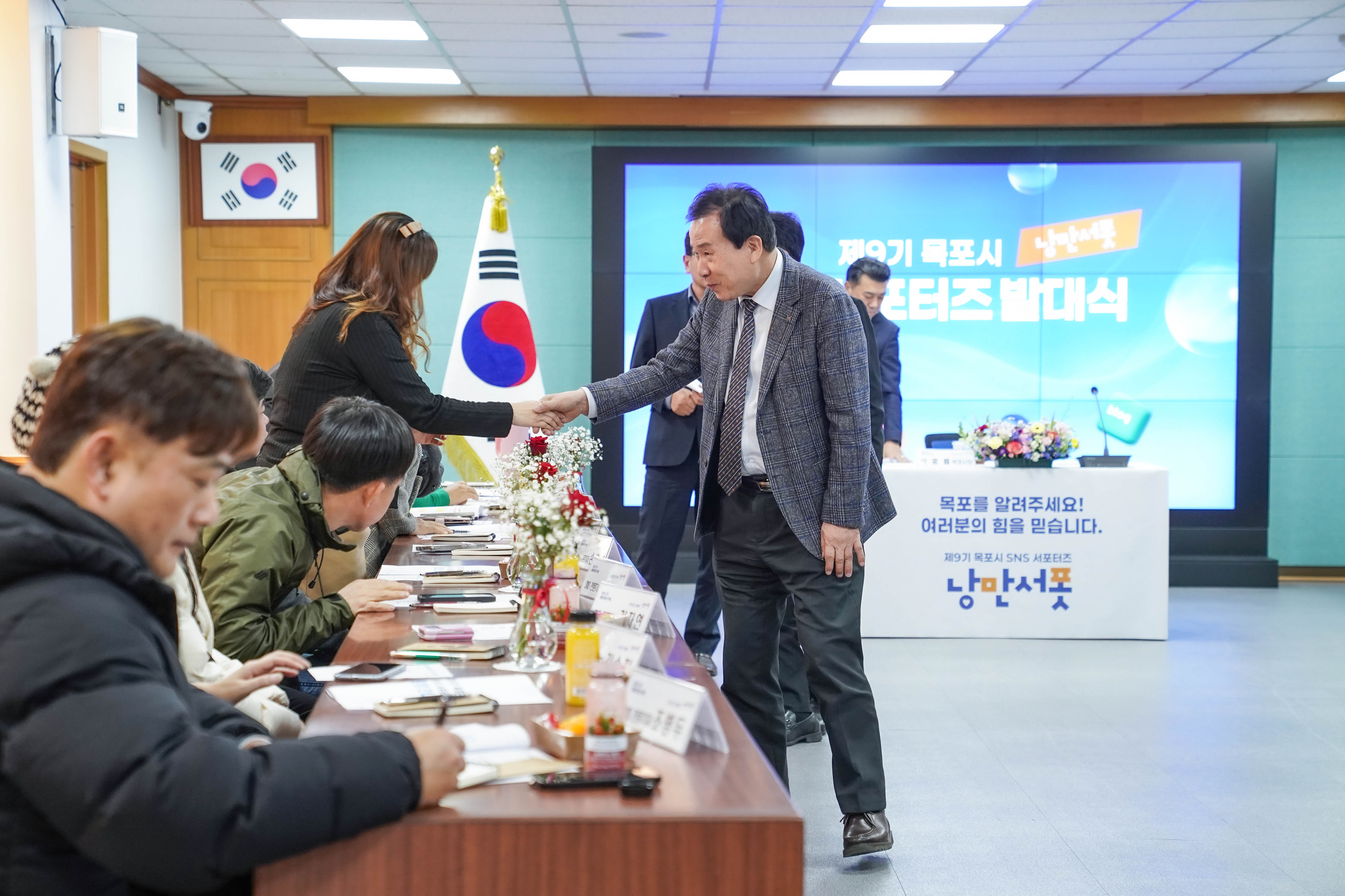 ‘제9기 목포시 낭만서포 SNS 서포터즈 발대식’ 행사 중, 양복을 입은 박홍률 목포시장이 참석자 중 한 명과 악수하고 있는 장면. 뒤쪽 대형 스크린에 “제9기 목포시 낭만서포 SNS 서포터즈 발대식”이라는 문구와 함께 SNS 아이콘(유튜브, 인스타그램, 블로그 등)이 배경으로 나타남. 책상 위에는 명찰과 꽃병이 놓여 있고, 한국 국기가 배경에 함께 있음.