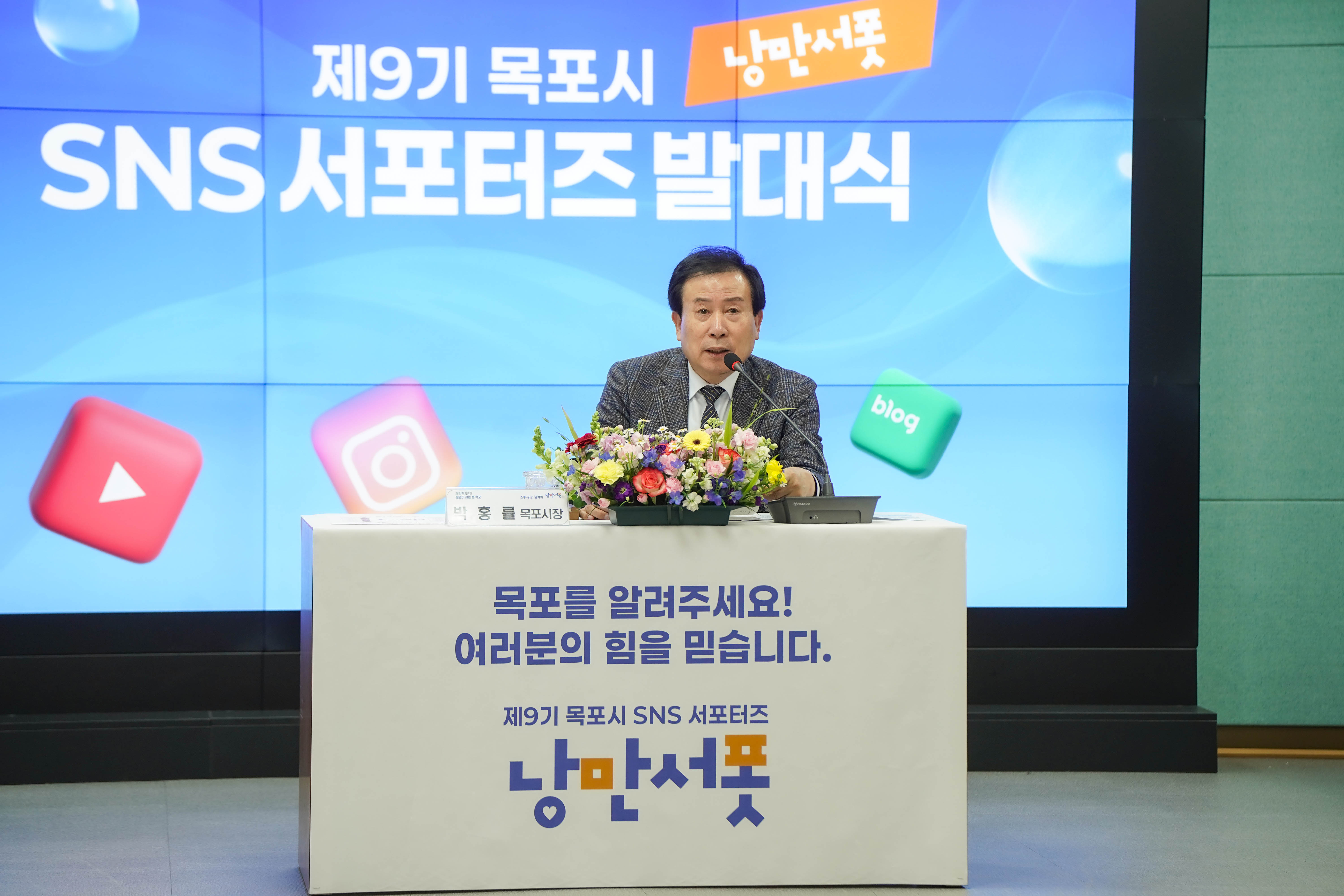 ‘제9기 목포시 낭만서포 SNS 서포터즈 발대식’에서 박홍률 목포시장이 연단에 앉아 마이크로 인사말을 전하는 모습. 연단 전면에는 다음과 같은 문구가 적혀 있음:
“목포를 알려주세요! 여러분의 힘을 믿습니다. 제9기 목포시 SNS 서포터즈 낭만서포.”
배경 스크린에는 같은 문구와 함께 ‘낭만서포’ 로고 및 유튜브, 인스타그램, 블로그 등의 SNS 아이콘이 떠 있음. 연단 위에는 형형색색의 꽃다발이 놓여 있음.