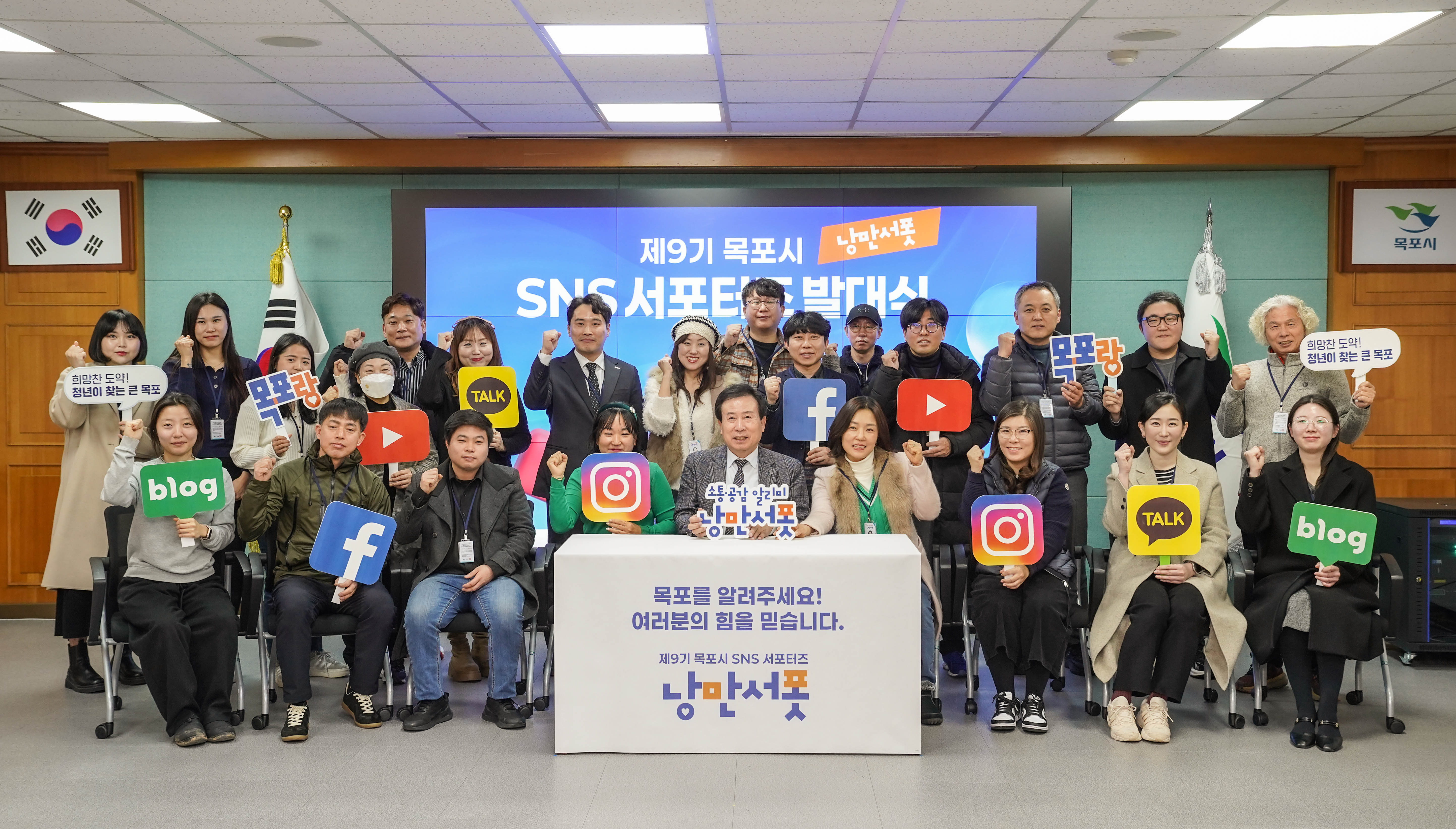‘제9기 목포시 낭만서포 SNS 서포터즈 발대식’ 단체 기념 촬영 사진. 가운데 박홍률 목포시장이 앉아 있으며, 양쪽으로 SNS 서포터즈들이 단체로 둘러서 있음. 각 인원은 인스타그램, 유튜브, 블로그, 페이스북, 카카오톡, ‘낭만서포’, ‘목포랑’, '희망찬 도약! 청년에 찾는 큰 목포' 등의 문구가 적힌 팻말을 들고 있음. 뒤쪽 배경 스크린에는 “제9기 목포시 낭만서포 SNS 서포터즈 발대식”이라는 문구가 큼직하게 표시되어 있음. 참석자 모두가 주먹을 쥐고 파이팅 포즈를 취하고 있음.