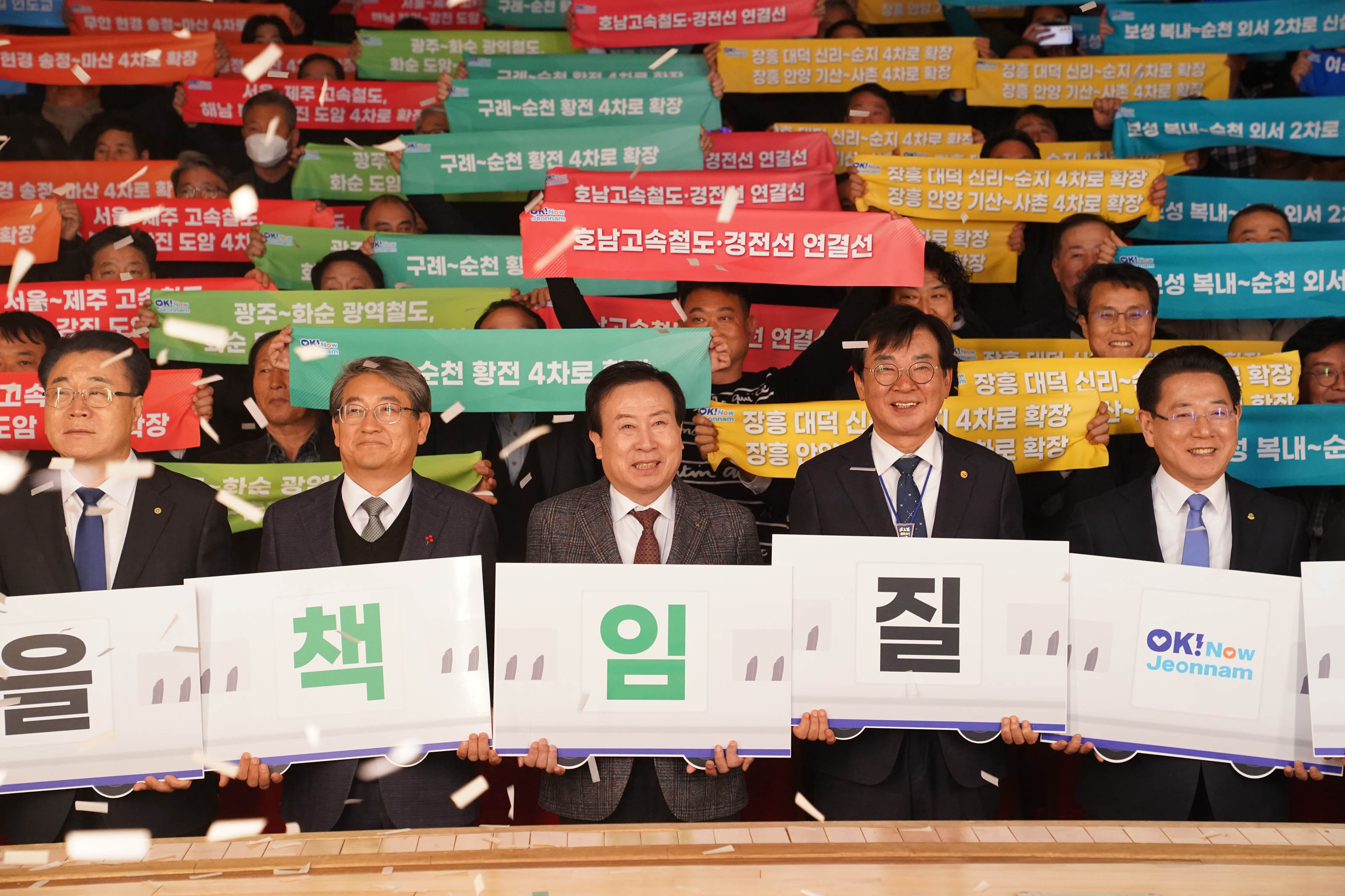 무대 앞줄에 박홍률 목포시장을 비롯한 다섯 명의 주요 인사들이 나란히 서서 각자 흰색 팻말을 들고 있는 모습. 팻말에는 차례대로 을, 책, 임, 질, OK! Now Jeonnam 이라고 적혀 있다. 뒤편 관중석에서는 여러 사람이 빨간색, 녹색, 노란색, 파란색 피켓을 들고 있다. 피켓에는 다음과 같은 문구가 적혀 있음:

호남고속철도·경전선 연결선

구례~순천 황전 4차로 확장

장흥 대덕 실리 4차로 확장 장흥 안양 기산~사촌 4차로 확장

광주~화순 광역철도, 화순 도암 고속도로

서울제주 고속철도, 광주보성 4차로 확장

서울~제주 고속철도, 전남 연장

장흥~안양 도암 고속도로

고흥(우주센터)~광주 고속도로

구례~순천 외서 2차로

장흥 대덕 실리~사촌 4차로 확장

서울~제주 고속철도, 제주 연장

함평 환경 중흥~마산 4차로 확장 등 다양한 교통 인프라 건설 관련 문구가 다채로운 색상의 피켓에 적혀 있음.