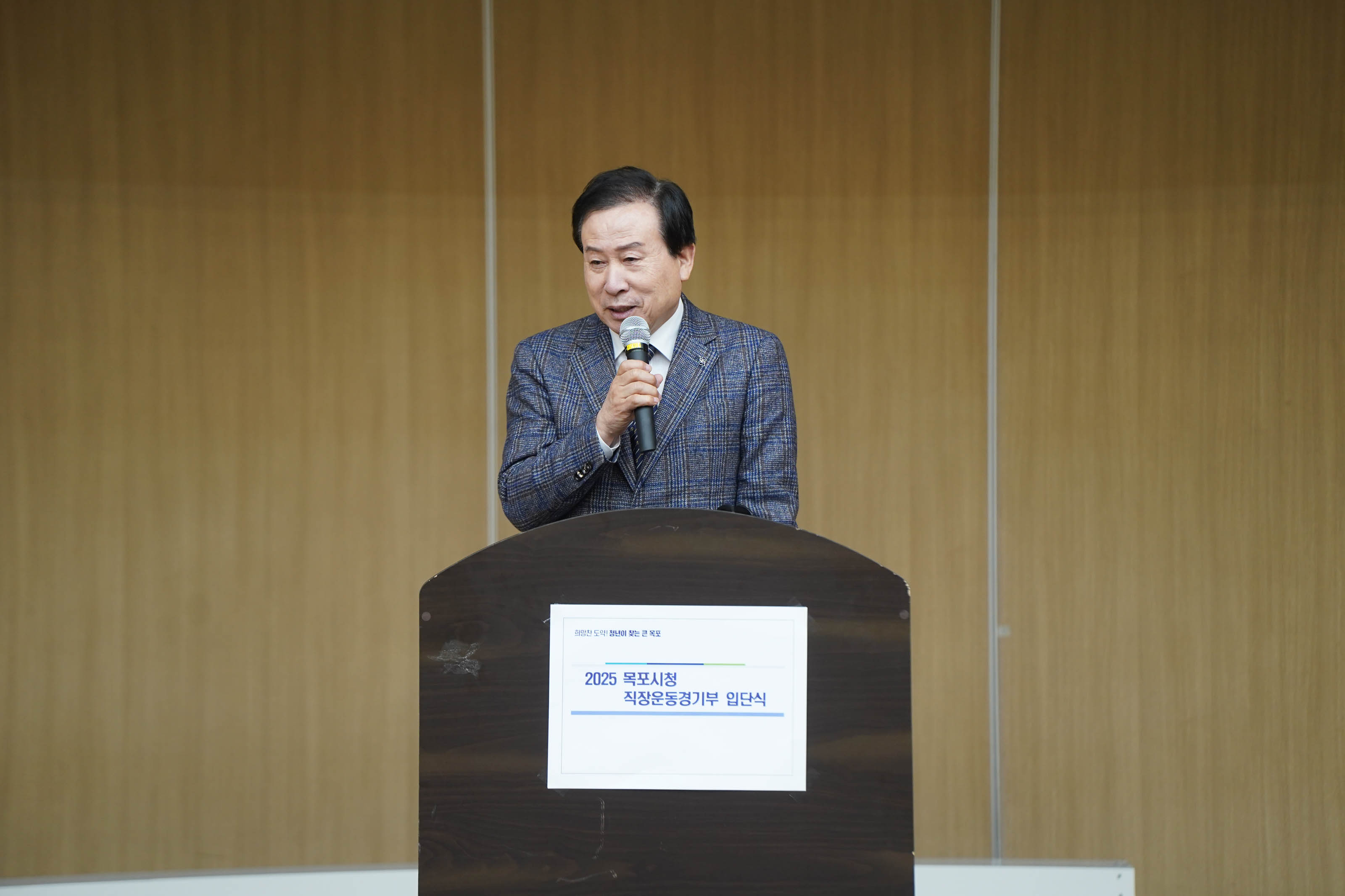 목포시청 직장운동경기부 입단식 행사에서 마이크를 들고 발언 중인 박홍률 목포시장의 클로즈업 모습. 연단에는 ‘2025 목포시청 직장운동경기부 입단식’이라는 텍스트가 적혀 있고, 뒤 배경은 나무색 벽으로 구성됨.