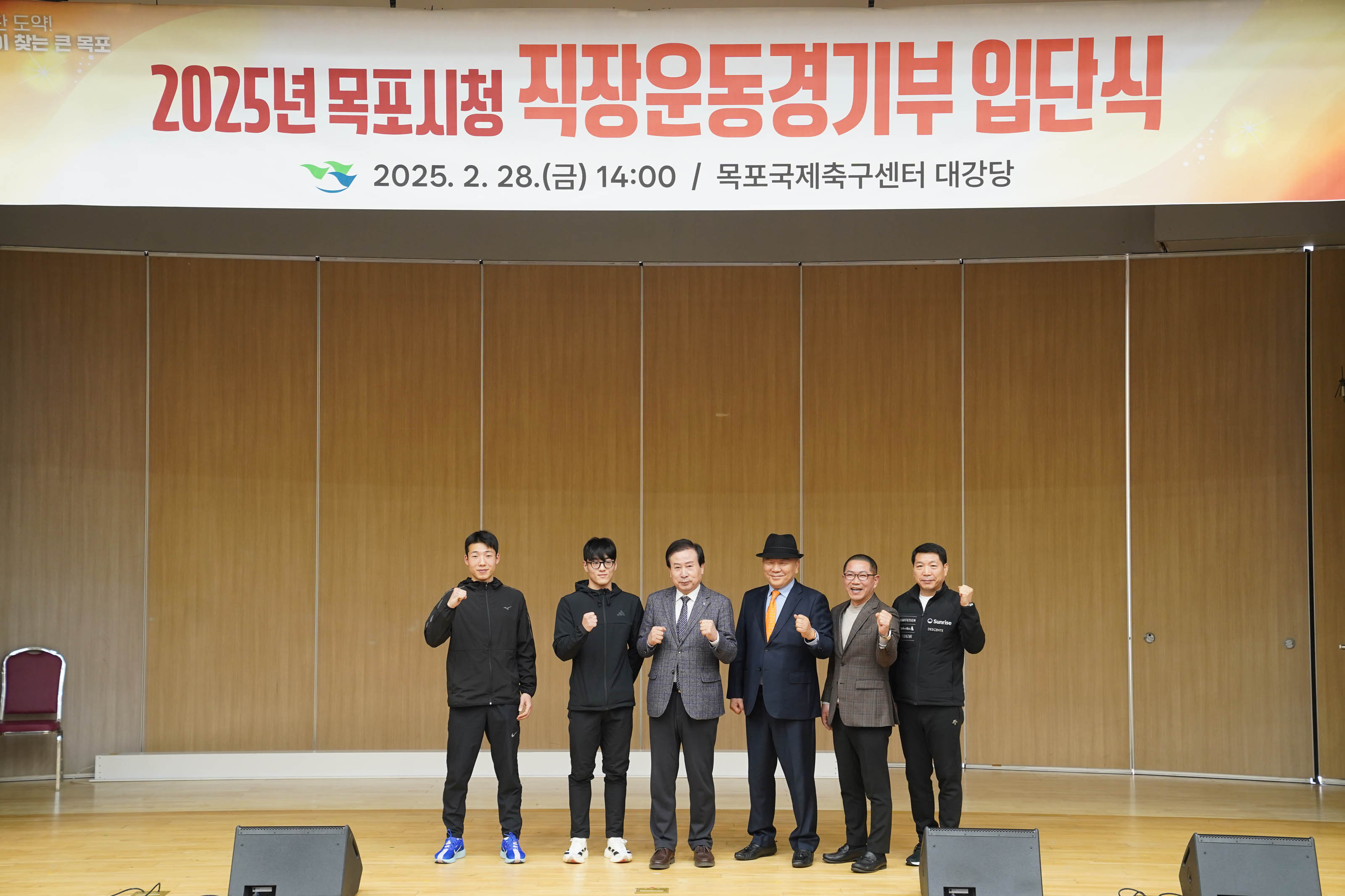 무대 중앙에서 박홍률 목포시장을 비롯한 여섯 명이 일렬로 서서 카메라를 바라보며 파이팅 포즈를 취하고 있다. 배경에는 ‘2025년 목포시청 직장운동경기부 입단식 2025.2.28(금) 14:00 / 목포국제축구센터 대강당’ 현수막과 행사 정보가 적혀 있고, 우측에는 태극기와 목포시 로고도 보인다.