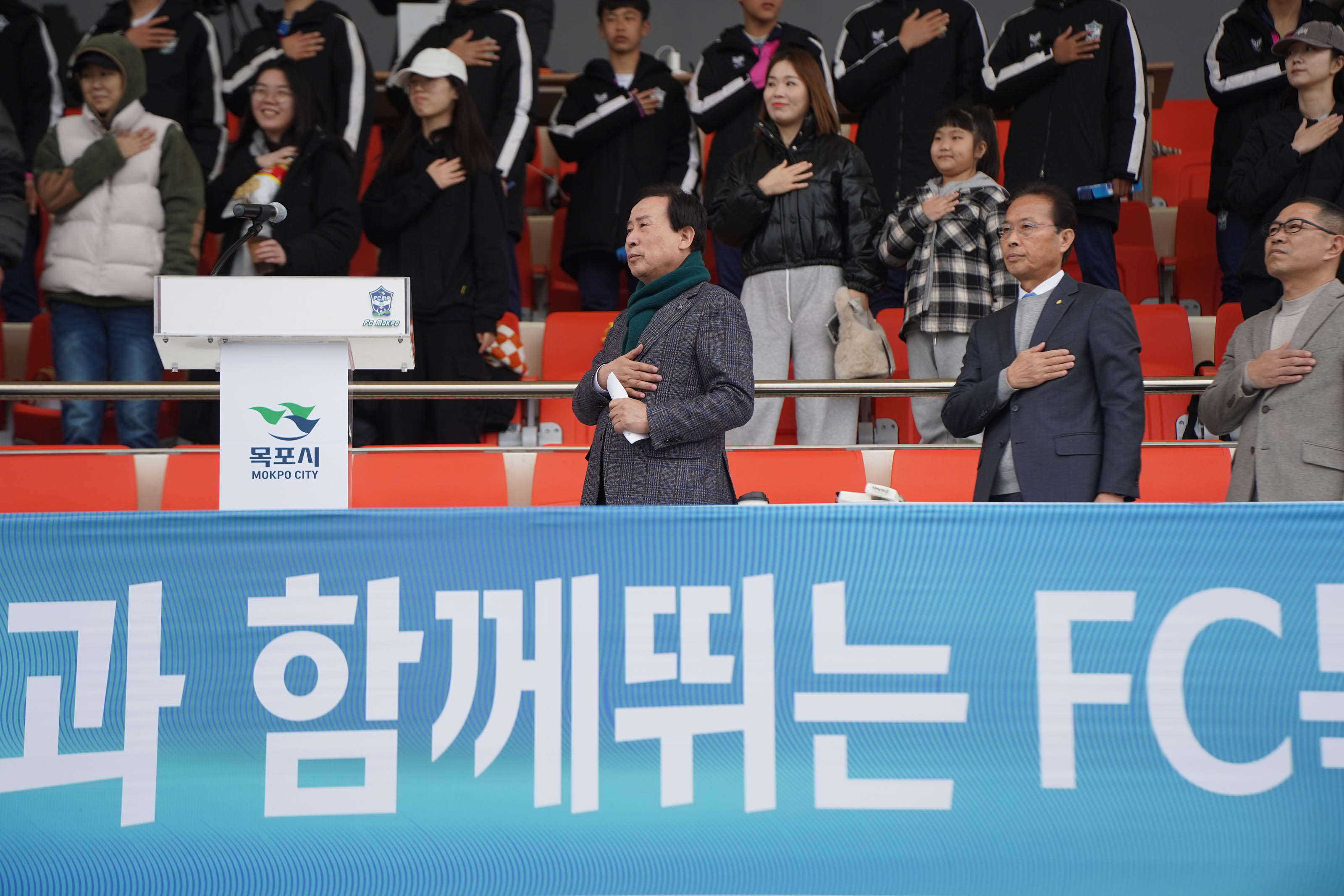 축구 경기장 스탠드에서 관중들과 내빈들이 국기에 대한 경례를 하고 있는 모습. 중앙 단상에는 '목포시 MOKPO CITY' 로고가 새겨진 연단이 설치되어 있고, 왼쪽에는 마이크가 놓여 있다. 앞줄에 있는 박홍률 목포시장과 두 명의 관계자들은 오른손을 왼쪽 가슴에 얹고 경례 중이며, 뒤쪽에는 다양한 연령대의 시민들과 젊은 학생들이 함께 일어서서 경례에 참여하고 있다.