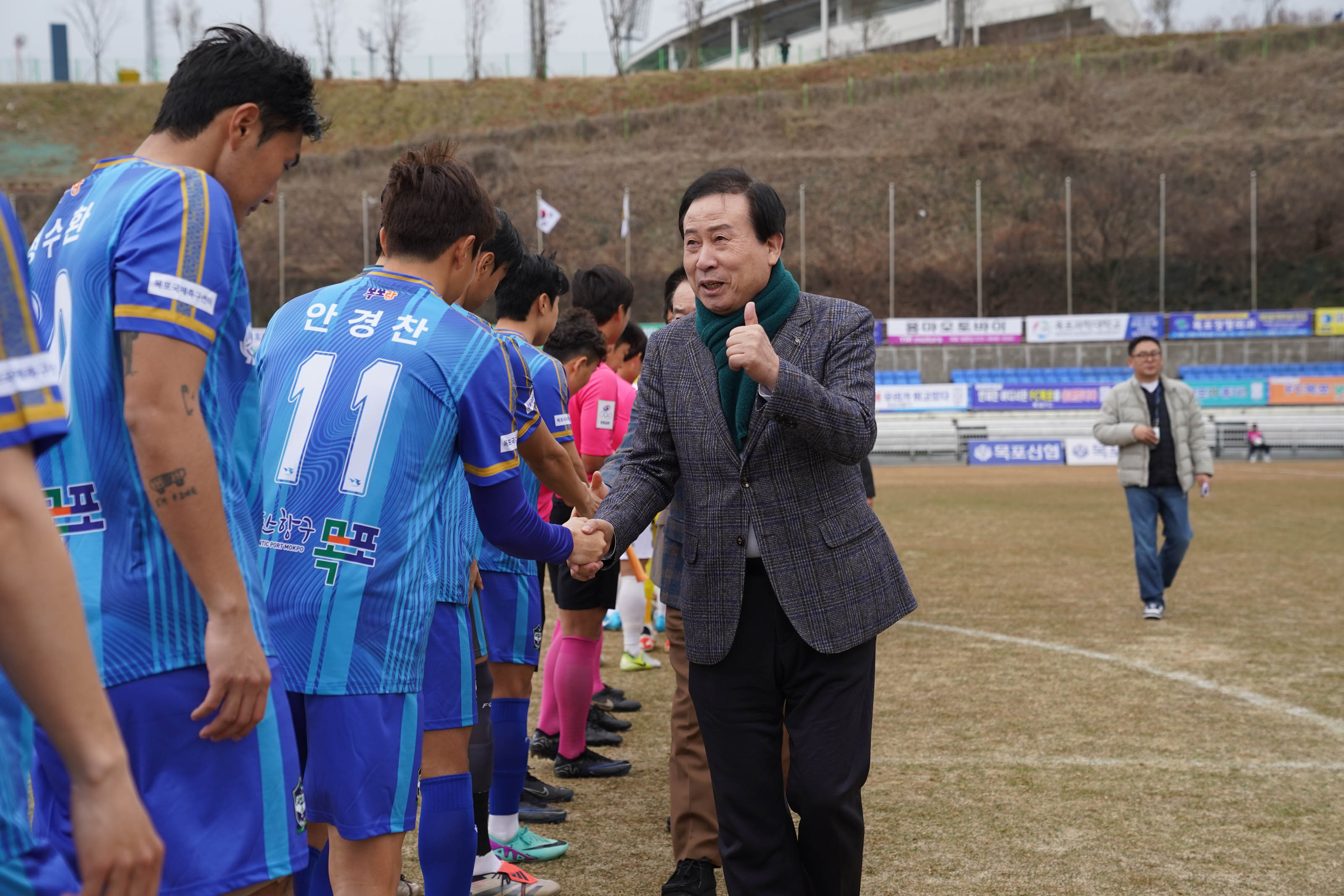 박홍률 목포시장이 파란색 유니폼을 입은 FC목포 선수들과 악수를 나누며 엄지를 들어 격려하고 있다. 선수들 유니폼에는 '안경찬' 등 글자가 적혀 있으며, 뒷편에는 광고 현수막들이 경기장 펜스를 따라 설치되어 있다. 주변에는 관계자들이 선수단 인사를 지켜보고 있다.