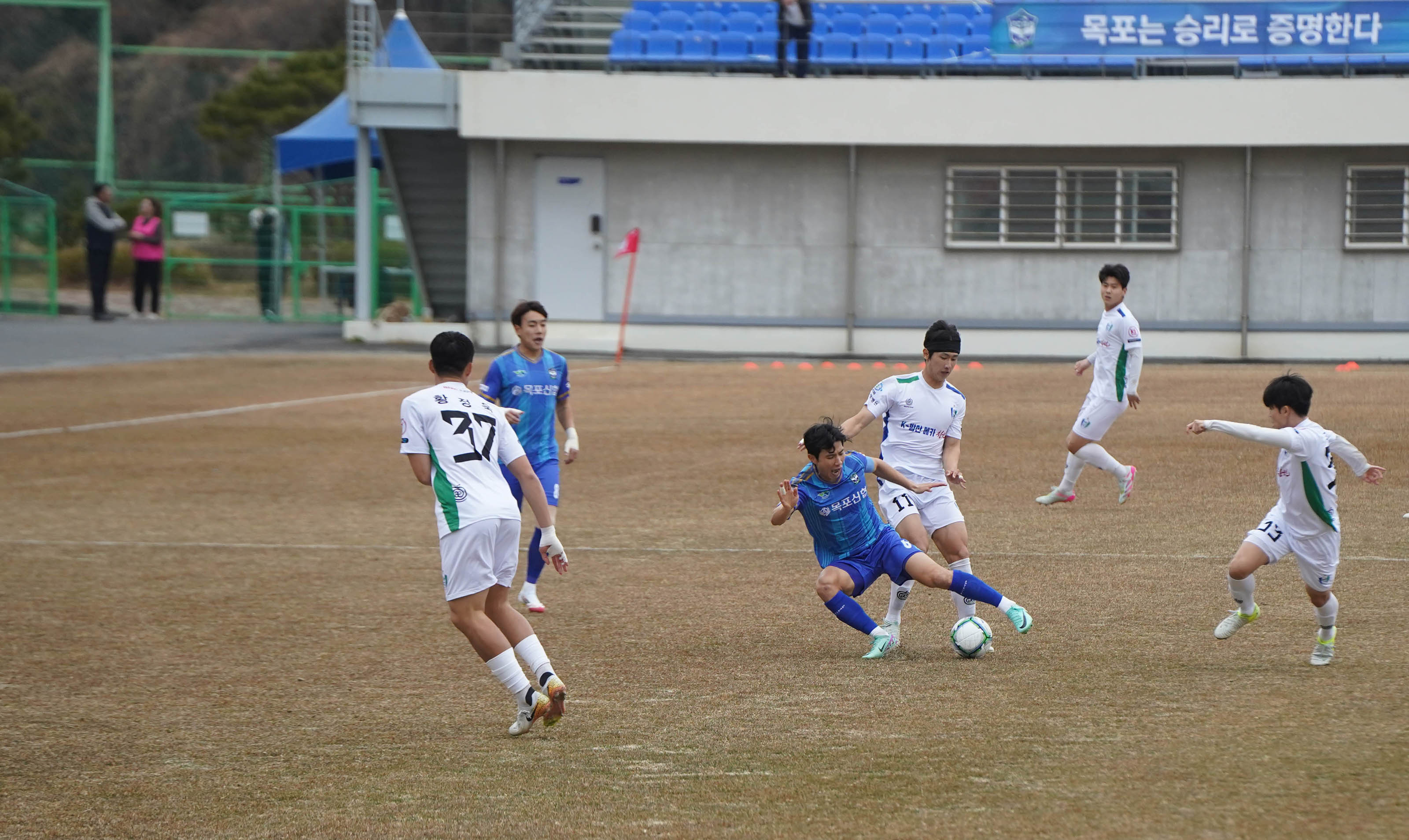 축구 경기 중 파란색 유니폼의 FC목포 선수 한 명이 흰색 유니폼의 상대 선수에게 태클을 거는 장면. 그 주위에는 다른 선수들이 달려오고 있으며, 뒤편 스탠드에는 파란색 좌석에 앉아 경기를 관람하는 관중들이 보인다.