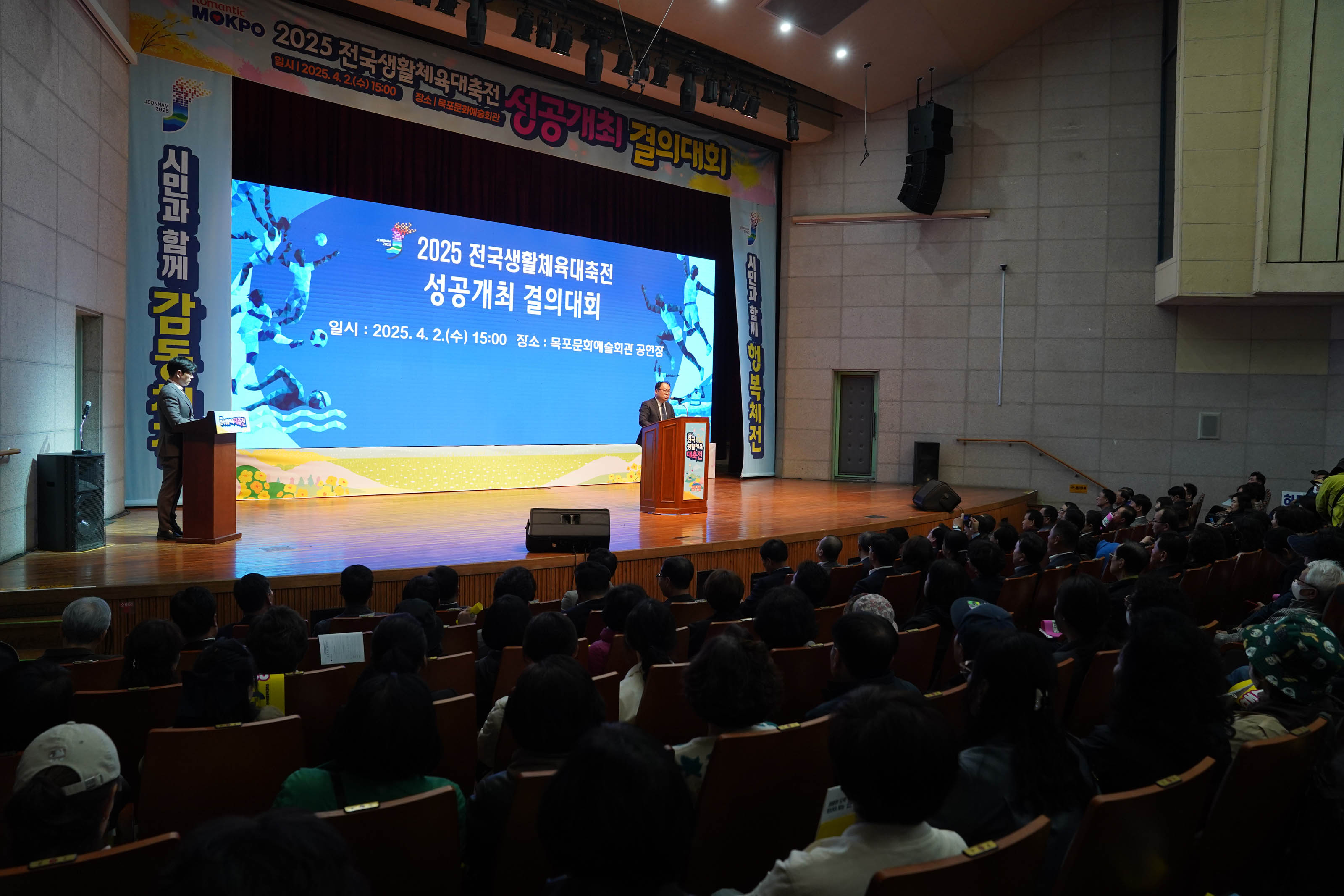 '2025전국생활체육대축전 성공개최 결의대회' 현수막이 위쪽에 설치되어 있고 '시민과 함께 감동체전', '시민과 함께 행복체전' 현수막이 스크린 양쪽으로 설치된 강당에서 '2025 전국생활체육대축전
성공개최 결의대회 일시:2025.4.2.(수) 15:00 장소: 목포문화예술회관 공연장' 문구가 적힌 스크린 앞에서 발표하는 관계자와 옆에 서있는 사회자 그리고 지켜보는 사람들의 모습