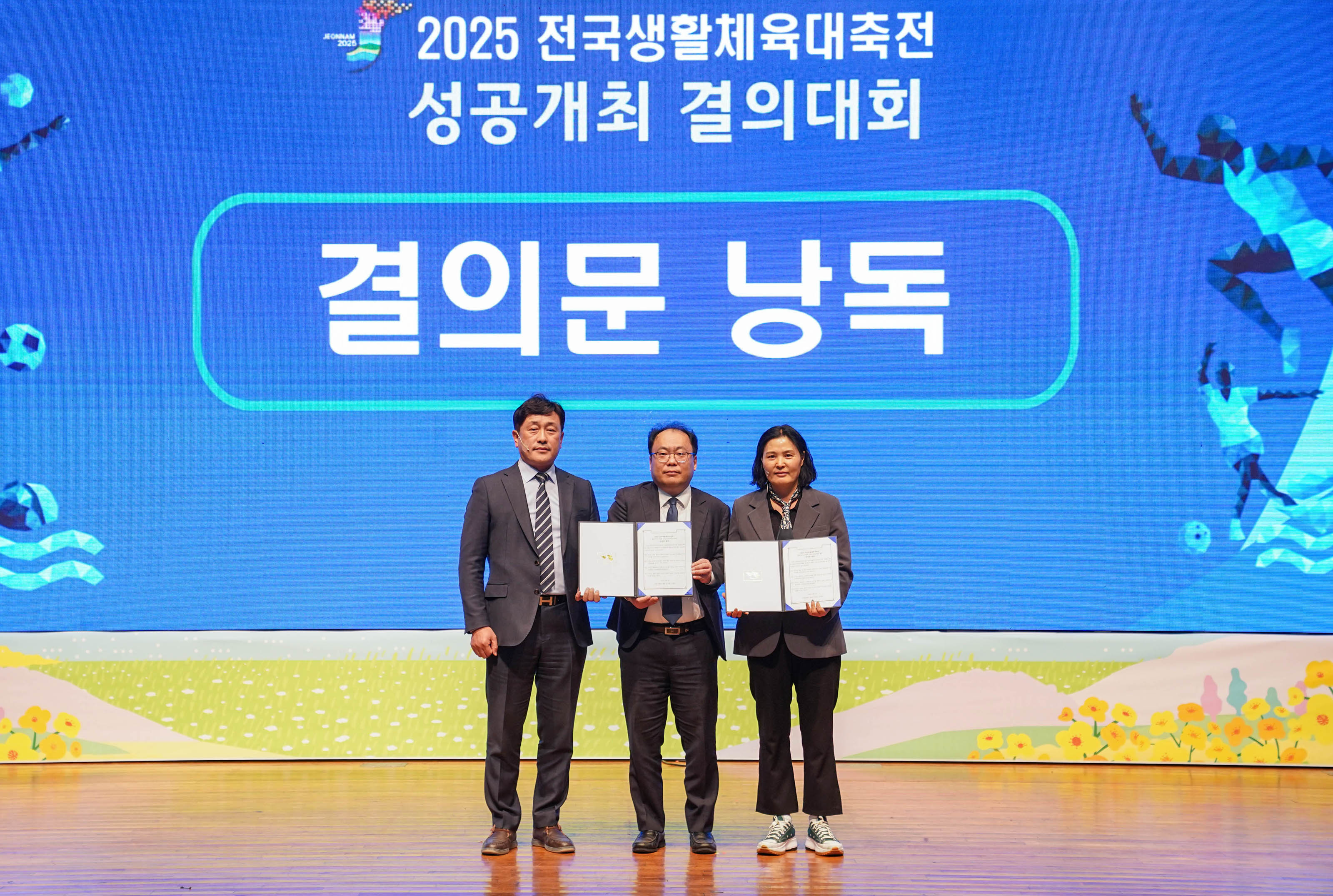 무대 중앙에서 세 명의 인물이 나란히 서서 결의문을 들고 기념 촬영을 하고 있다. 배경 스크린에는 '2025 전국생활체육대축전 성공개최 결의대회' 라는 문구와 함께, '결의문 낭독' 이라는 큰 글씨가 표시되어 있다. 스크린 좌우에는 다양한 스포츠 활동을 형상화한 그래픽 이미지가 그려져 있으며, 무대 아래에는 꽃과 들판이 그려진 일러스트 배경이 보인다.