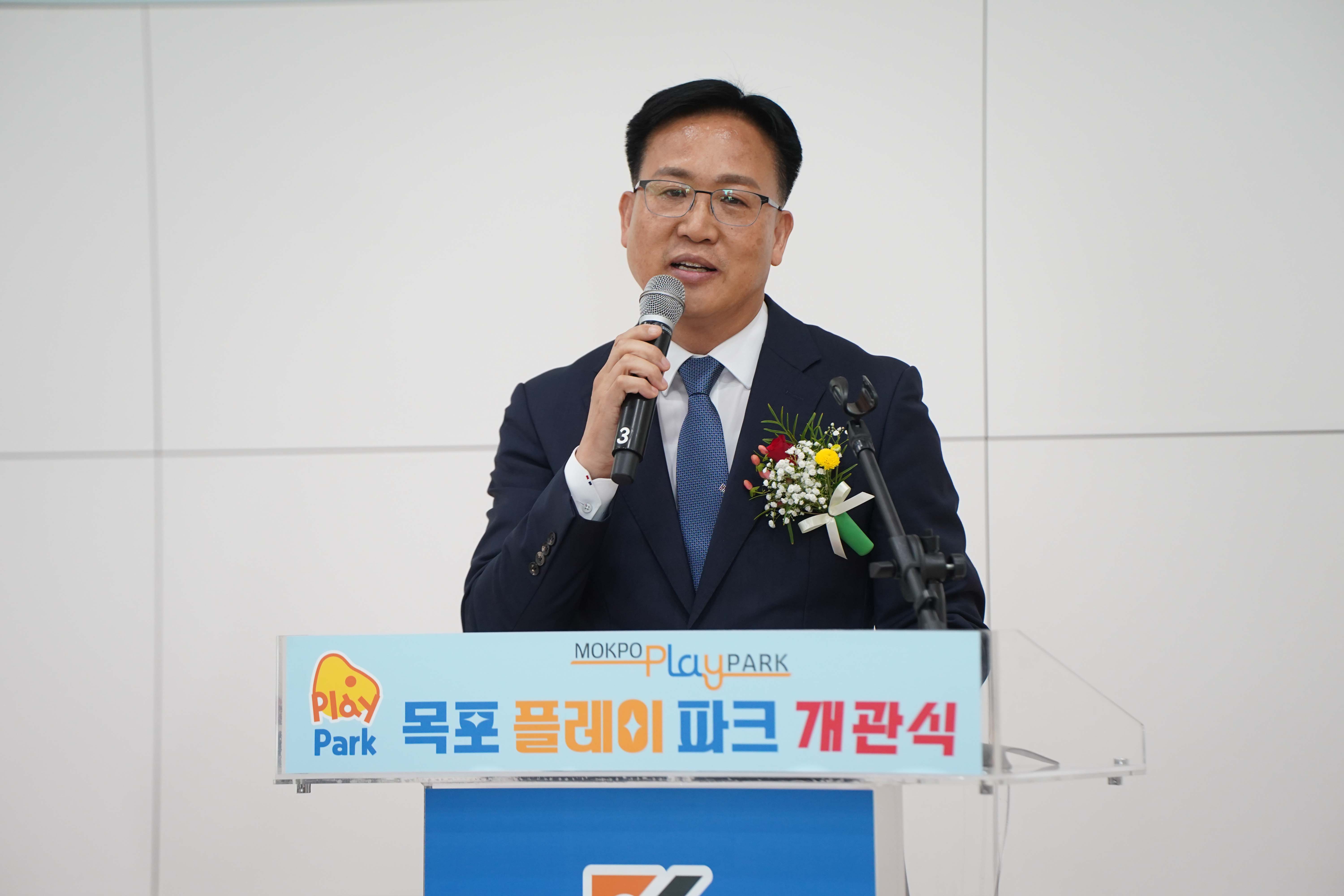 'MOKPO PLAY PARK 목포 플레이 파크 개관식' 단상에서 마이크를 잡고 발언 중인 내빈의 상반신 모습.
