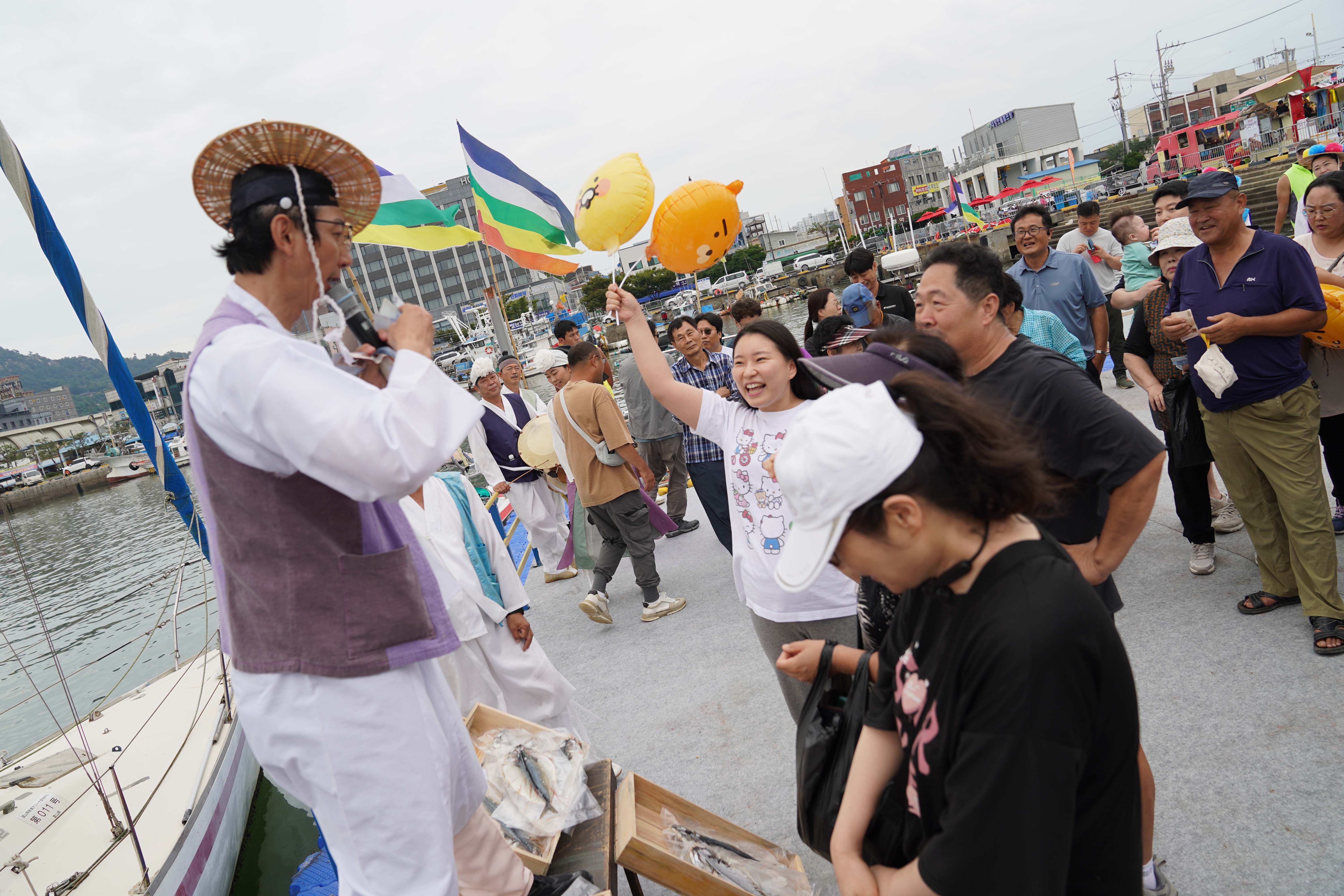 목포항구축제에서 삿갓을 쓰고 전통 의상을 입은 경매사가 배 위에서 마이크를 잡고 수산물 파시(波市) 경매를 진행하는 모습. 한 여성이 라이언 풍선을 들고 밝게 웃고 있으며, 주변으로 많은 관람객이 경매를 구경하고 있다.