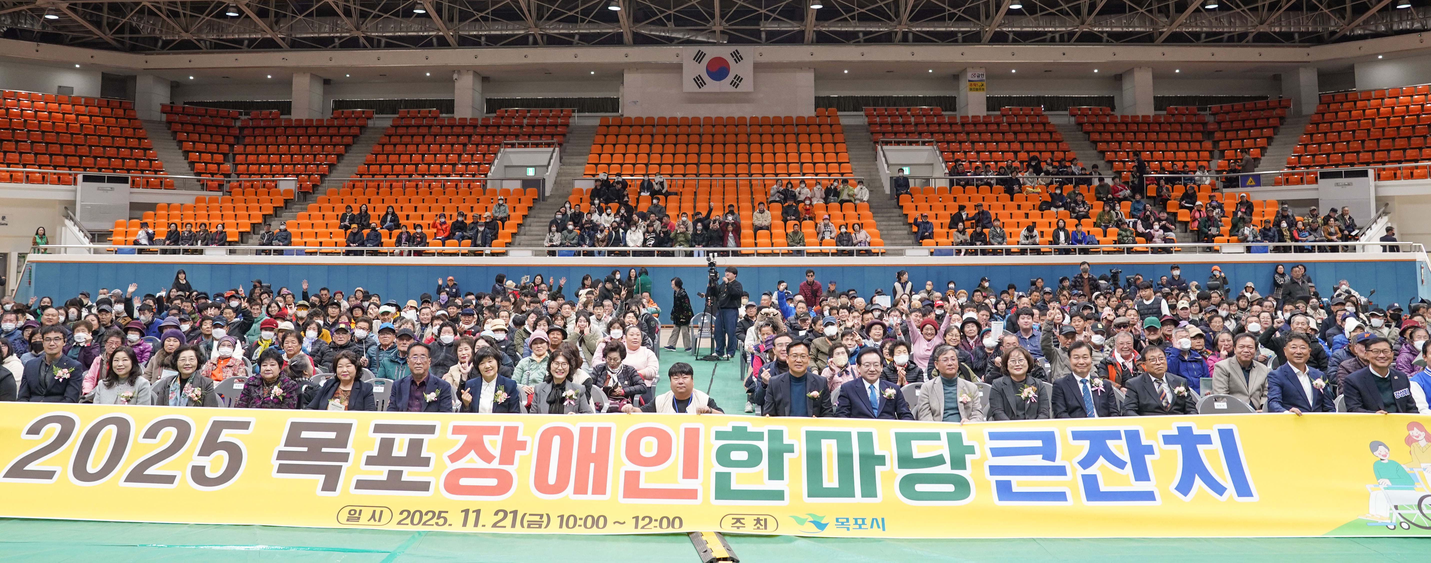 실내 체육관을 가득 채운 행사 참가자들의 단체 사진. 앞줄 내빈들이 '2025 목포장애인 한마당큰잔치, 일시 2025. 11. 21(금) 10:00~12:00, 주최 목포시'라고 적힌 대형 노란색 현수막을 펼쳐 들고 있으며, 관객석의 시민들이 손을 흔들며 환호하고 있음.