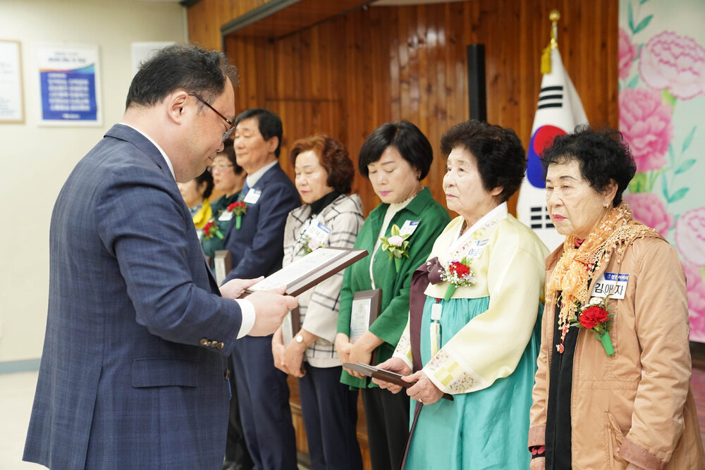 정장을 입고 안경을 낀 남성이 주황빛 점퍼를 입은 여성에게 패를 수여하고 있다