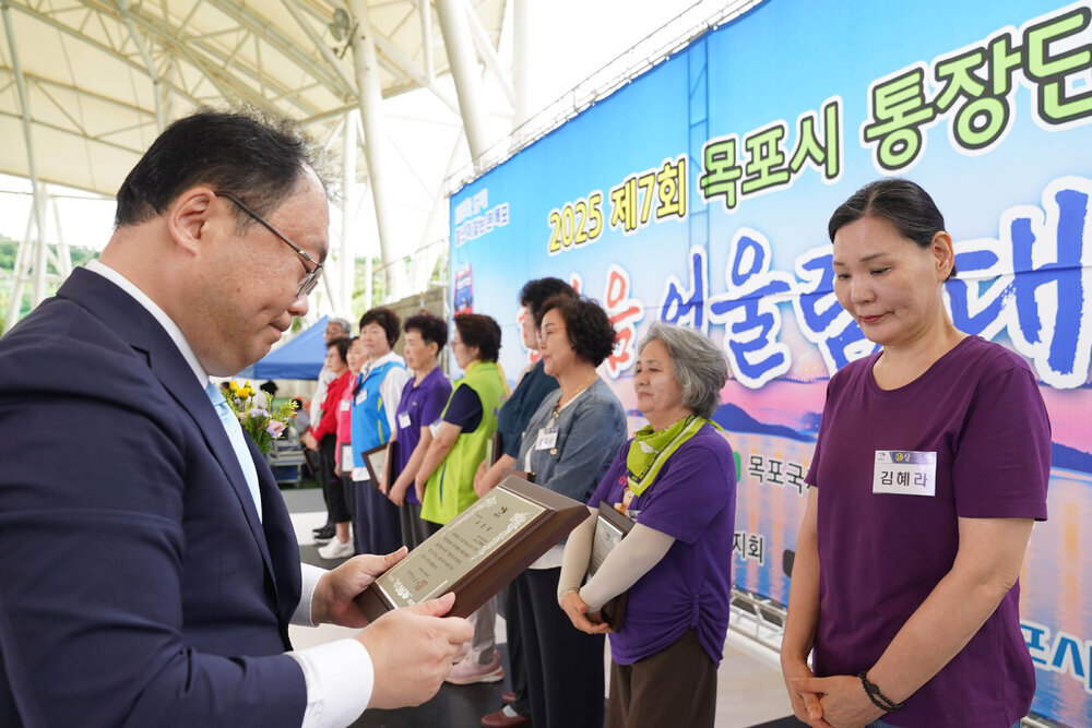정장을 입고 안경을 낀 남성이 보라색 반팔티를 입은 김혜라에게 패를 수여하고 있다