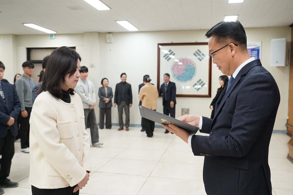 2026년 상반기 4급이하 정기인사로 정장을 입은 남성이 실내 행사장에서 한 여성에게 상장을 전달하며 내용을 읽고 있고, 여성은 흰색 트위드 자켓을 입고 두 손을 모은 상태로 서 있으며 뒤쪽으로 참석자들이 줄지어 서서 행사를 지켜보고 있다.