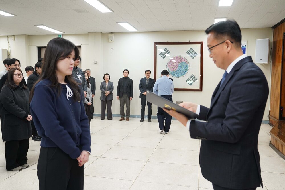 2026년 상반기 4급이하 정기인사로 정장을 입고 안경을 쓴 남성이 실내 행사장에서 한 여성에게 상장을 전달하며 내용을 읽고 있고, 여성은 남색 가디건을 입고 두 손을 모은 채 서 있으며 뒤쪽에는 참석자들이 줄지어 서서 행사를 지켜보고 있다.