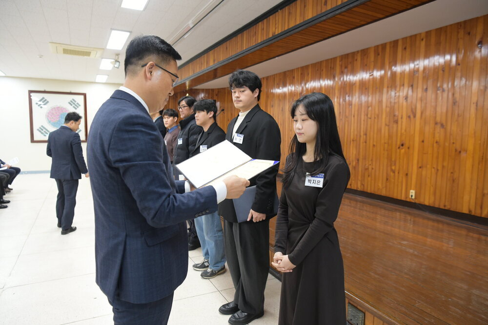 3월 정례조회에 정장을 입고 안경을 낀 남성이 검은색 원피스를 입은 박지은에게 패를 수여하고 있다.