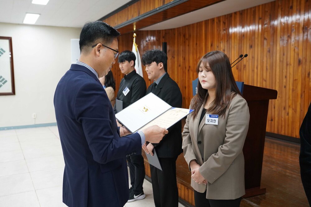 4월 정례조회에 정장을 입고 안경을 낀 남성이 짙은 오트밀색 자켓을 입은 오지인에게 패를 수여하고 있다